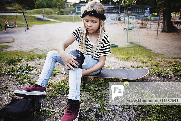 Girl tying kneepad while sitting on skateboard at park