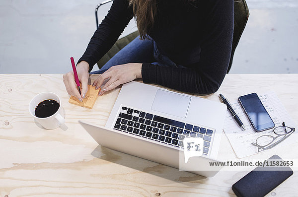 Hochwinkelansicht der jungen Frau beim Schreiben auf Klebezettel per Laptop am Schreibtisch