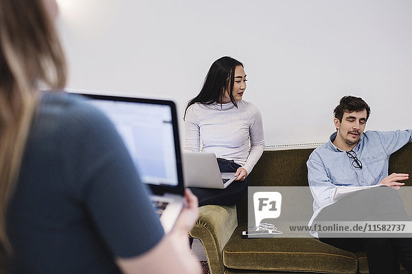 Young woman looking at male colleague sitting on sofa at office