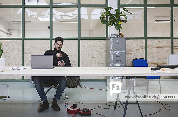 Junger Mann  der Musik über das Smartphone hört  während er mit dem Laptop am Schreibtisch im Büro sitzt.