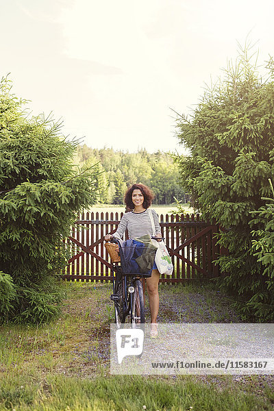 Volle Länge der Frau mit Fahrrad am Zaun stehend inmitten von Pflanzen