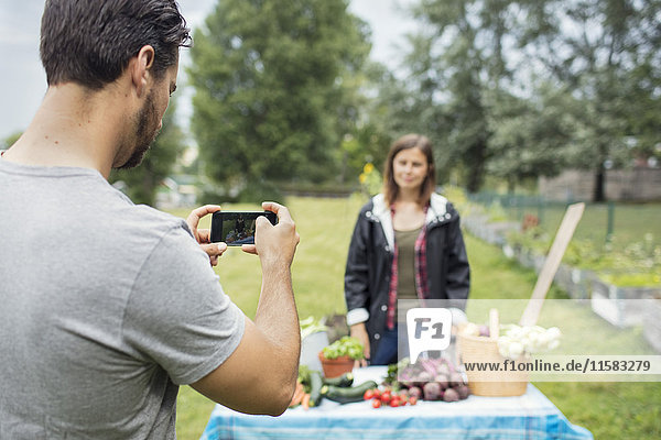 Mittlerer Erwachsener Mann  der eine Frau fotografiert  die im Stadtgarten neben frisch geerntetem Gemüse steht.