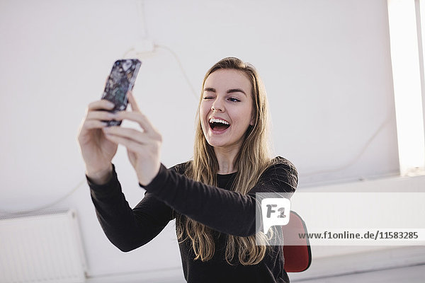 Fröhliche weibliche Bloggerin blinzelt  während sie Selfie durch Smartphone im Büro nimmt.