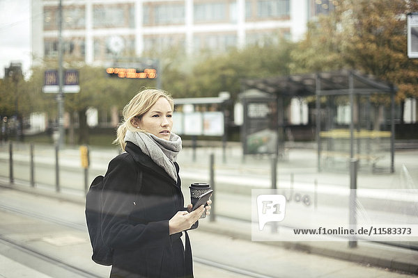 Mittlere erwachsene Geschäftsfrau mit Handy und Einweg-Kaffeetasse an der Straßenbahnhaltestelle