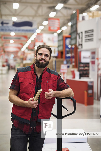 Portrait of confident salesman holding folding ruler while standing by handtruck in hardware store