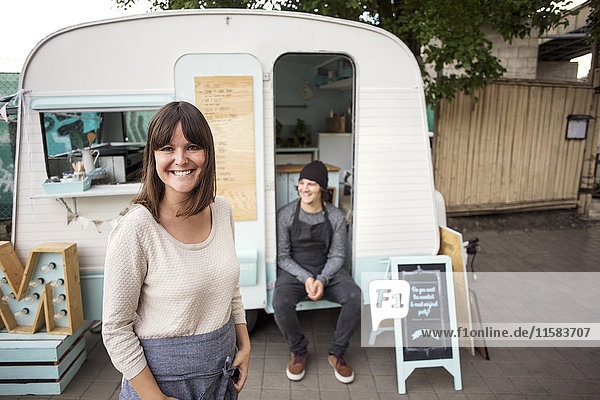 Portrait of happy female owner standing on street while coworker sitting in food truck