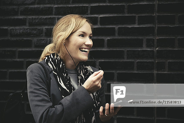 Smiling mid adult woman using hands-free device against brick wall