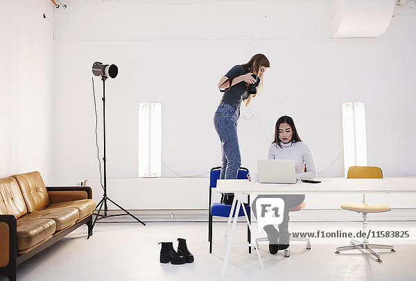 Blogger photographing female colleague using laptop at desk in creative office