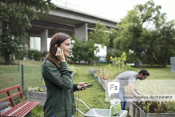 Mid adult woman talking on mobile phone with man planting in background at urban garden