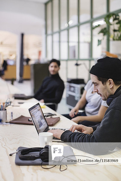 Junger Mann mit Laptop am Schreibtisch und Kollegen im Hintergrund