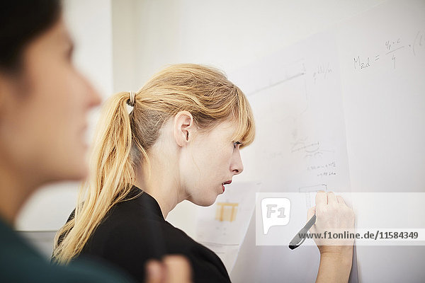 Seitenansicht der mittleren erwachsenen Geschäftsfrau auf Whiteboard mit Kollegin im Vordergrund im Büro