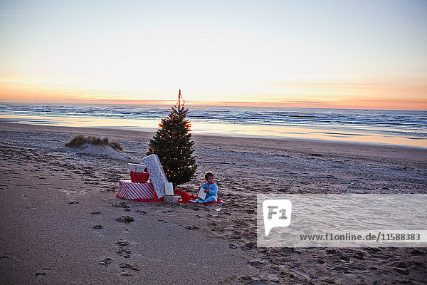 Junges Mädchen sitzt am Strand mit Weihnachtsbaum und Geschenken