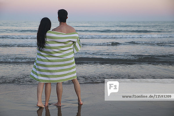 Back view of young couple on the beach wrapped together in towel watching sunset