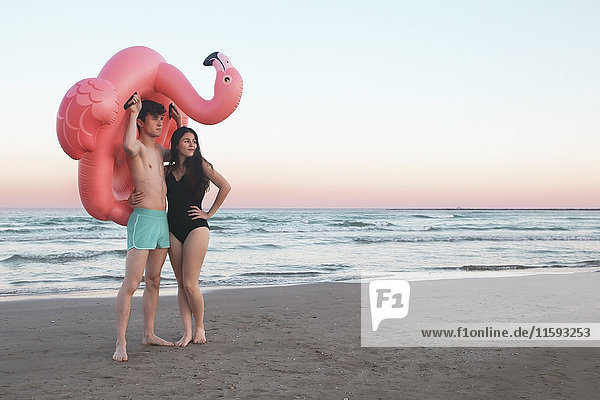 Junges Paar mit aufblasbarem rosa Flamingo am Strand bei Sonnenuntergang