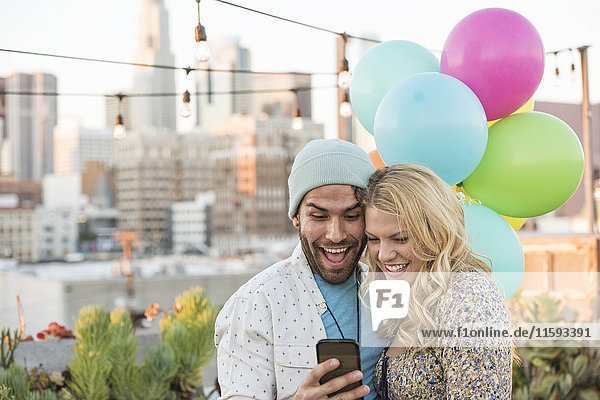 Junges Paar mit Luftballons auf der Dachterrasse mit Smartphone