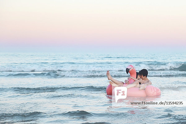 Verliebtes Paar schwebt bei Sonnenuntergang mit aufblasbarem rosa Flamingo auf dem Meer.