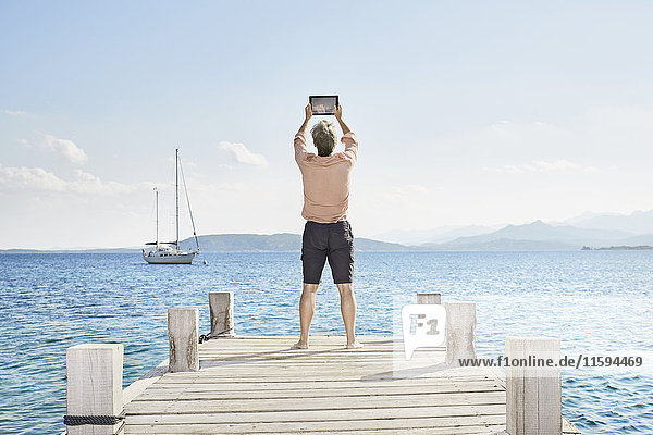 Back view of man standing on jetty taking selfie with tablet