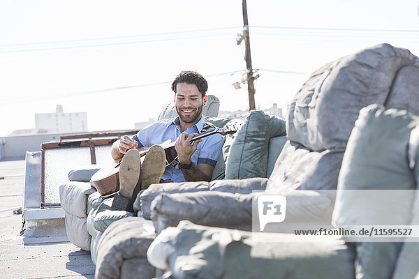 Young man on rooftop sitting on sofa and playing guitar