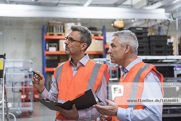 Two colleagues in factory hall wearing safety vests holding clipboard