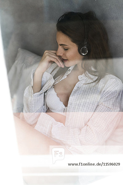 Young woman behind windowpane listening music with headphones at home