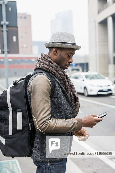 Stilvoller Mann mit Rucksack neben der Straße und Blick aufs Handy