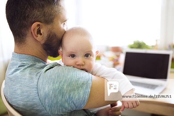 Father holding baby son at home with laptop on table