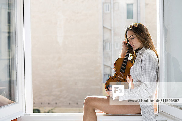 Young woman with eyes closed sitting on window sill holding violin