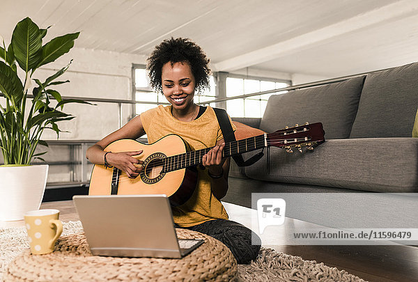 Lächelnde junge Frau zu Hause beim Gitarrespielen auf dem Laptop