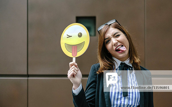 Geschäftsfrau hält Emoji lächelnd mit herausstehender Zunge in der Hand