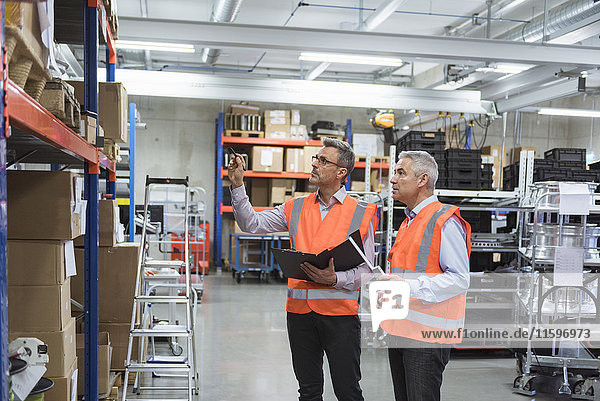 Two colleagues in factory hall wearing safety vests holding clipboard