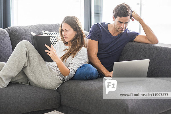 Young couple on couch relaxing with book and laptop
