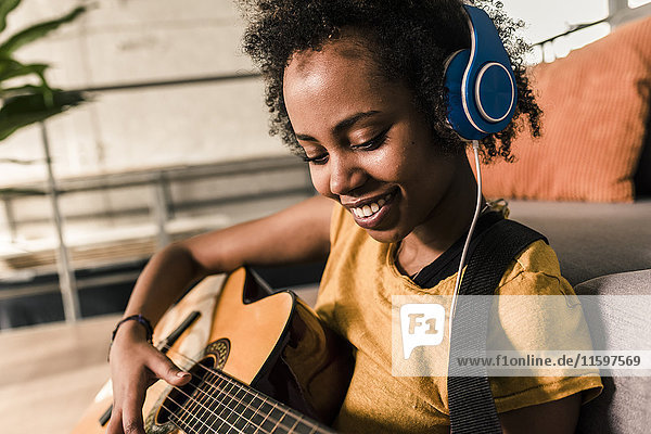 Lächelnde junge Frau zu Hause mit Kopfhörern beim Gitarrespielen