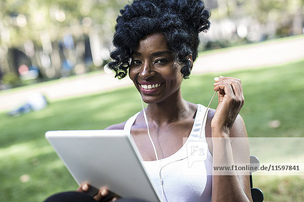 Portrait of smiling woman sitting in a park using tablet and earphones