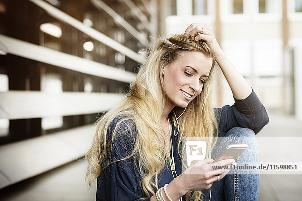 Porträt einer lächelnden blonden jungen Frau mit Kopfhörern beim Blick aufs Handy