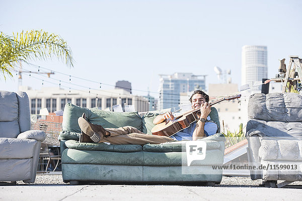 Junger Mann auf dem Dach auf dem Sofa liegend und Gitarre spielend