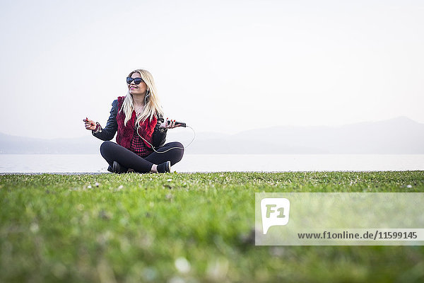 Smiling blond woman sitting on a meadow listening music with smartphone and earphones