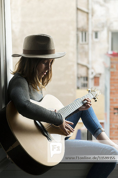 Junge Frau sitzt im Fensterrahmen und spielt Gitarre