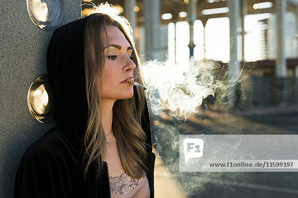 Porträt einer jungen Frau in Kapuzenjacke mit Zigarette im Gegenlicht