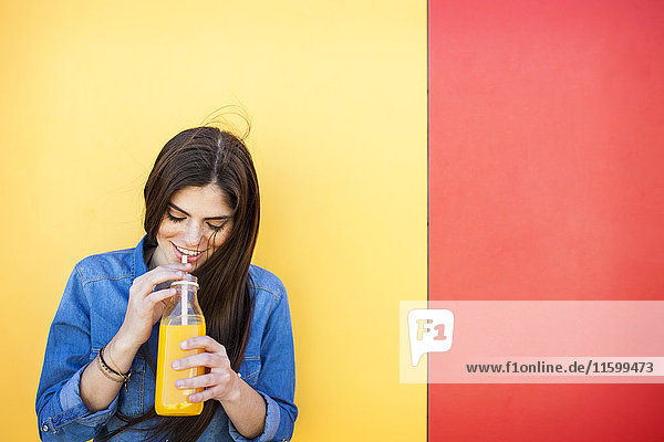 Lächelnde junge Frau vor bunter Wand  die Orangensaft trinkt.