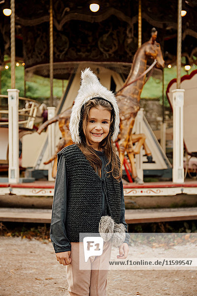 Portrait of smiling little girl wearing fur hat standing in front of children's carousel