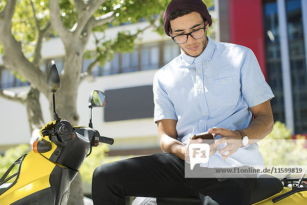 Junger Mann sitzt auf einem Motorroller und benutzt ein Handy.