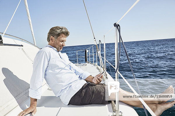 Erwachsener Mann sitzt auf seinem Segelboot und schaut auf sein Handy.
