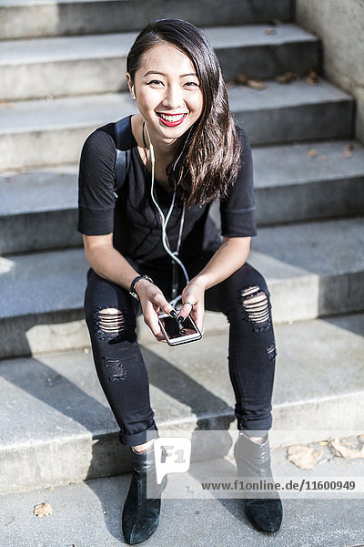 Portrait of young woman dressed in black sitting on steps listening music with earphones and cell phone