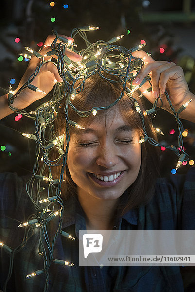 Mixed Race woman holding string lights on head near Christmas tree