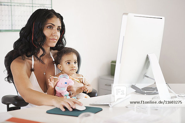 Gemischtrassige Mutter benutzt Computer und hält ihre kleine Tochter