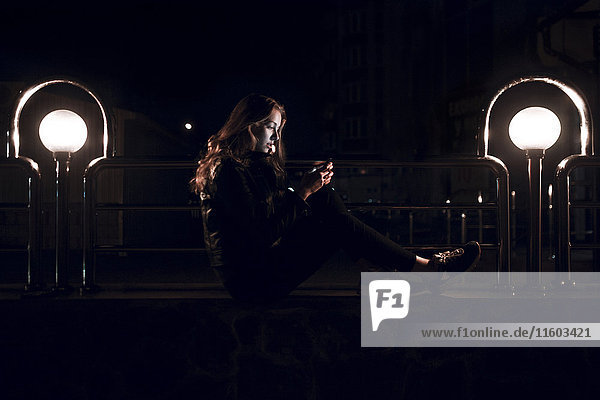 Kaukasisches Teenager-Mädchen sitzt nachts auf einer Bank und schreibt eine SMS auf einem Handy