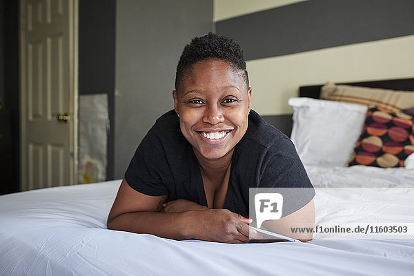 Lächelnde schwarze Frau  die auf dem Bett liegt und ein Handy hält