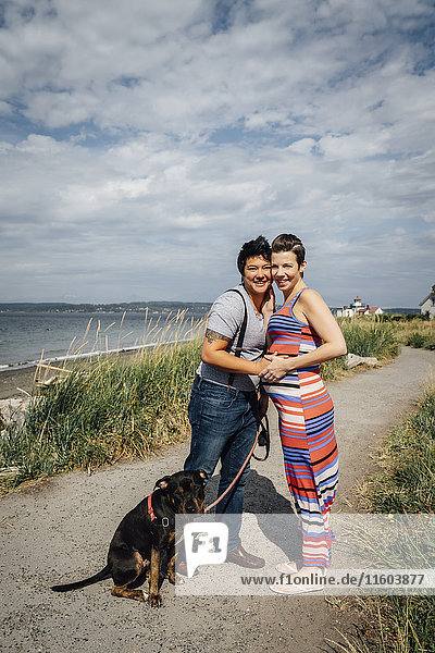 Porträt eines schwangeren lesbischen Paares mit Hund  das sich am Wasser umarmt