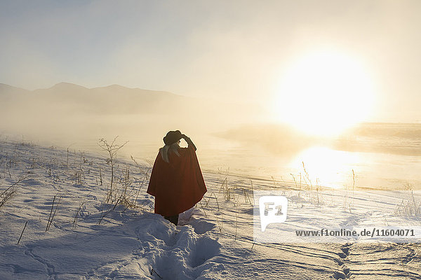 Kaukasische Frau  die ihre Augen in einer Winterlandschaft bei Sonnenuntergang abschirmt