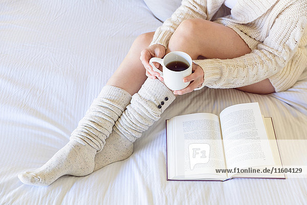 Caucasian woman reading book on bed and drinking coffee